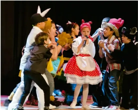Fotografía de niños y niñas representando una obra de teatro escolar en el escenario: una niña con vestido rojo de lunares y mandil sostiene un micrófono mientras los demás actúan a su alrededor con disfraces.