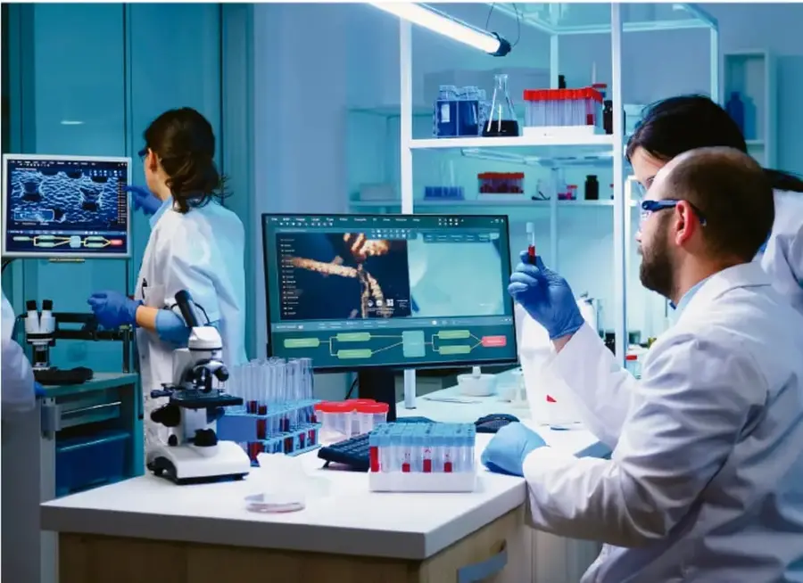 Fotografía de un laboratorio tecnológico con tres personas de bata blanca trabajando: una frente a un monitor con simulación, otra observando un microscopio y un hombre manipulando un tubo de muestra, rodeados de equipo científico y computadoras.
