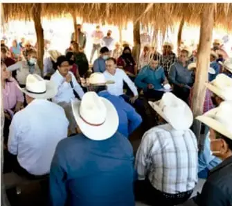 Reunión comunitaria en un espacio con techo de palma, personas sentadas en círculo, algunas con sombreros, dialogando.