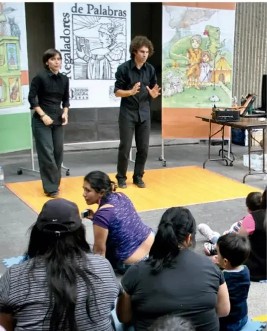 Fotografía de una sesión de narración oral al aire libre: dos narradores de pie frente a un público de niños y adultos sentados, con la cartelera "Regaladores de Palabras" y murales de cuentos de fondo.