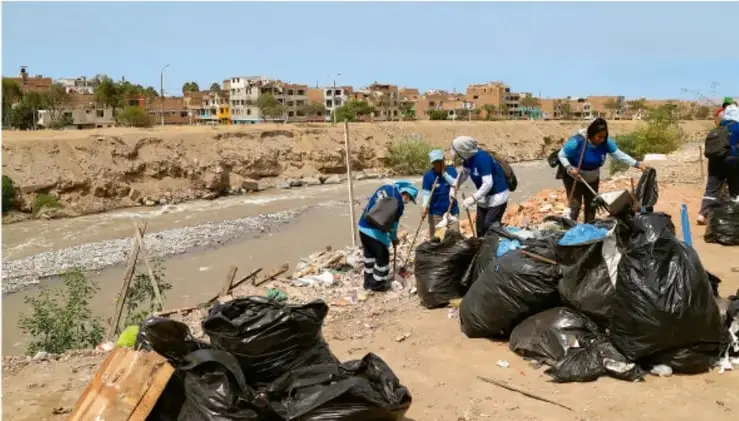 Fotografía de estudiantes con uniformes y guantes recogiendo basura en bolsas negras en la ribera de un río, con cerros y viviendas al fondo, mostrando una acción de limpieza comunitaria.