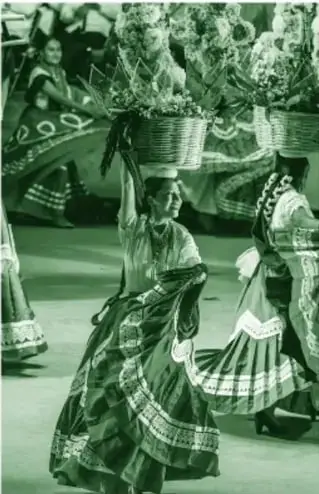 Fotografía a color (en tinta verde) de tres o cuatro mujeres bailando en la Guelaguetza, con faldas amplias, trenzas con listones y cestos de flores sobre la cabeza; al fondo se distingue el escenario.