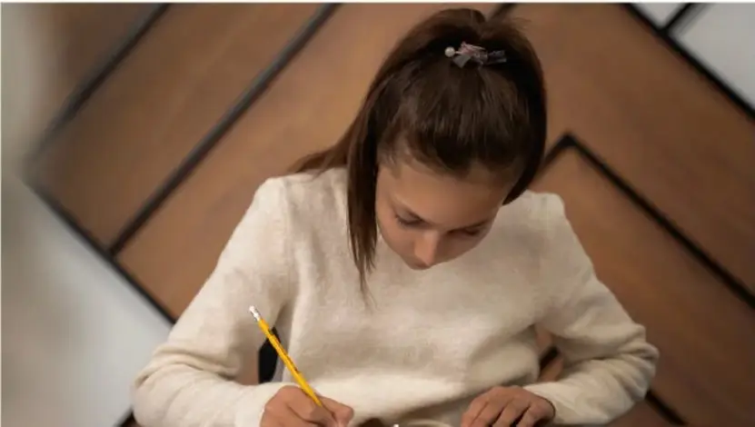Fotografía de una estudiante escribiendo con un lápiz en un cuaderno, concentrada, sobre una mesa con piso de madera.