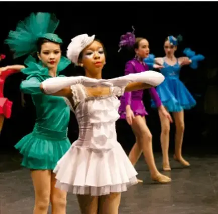 Fotografía de cinco bailarinas en escenario con vestidos de colores (verde, blanco, rosa, morado, azul) y tocados de plumas, en pose de danza, ilustrando el tema del performance artístico.