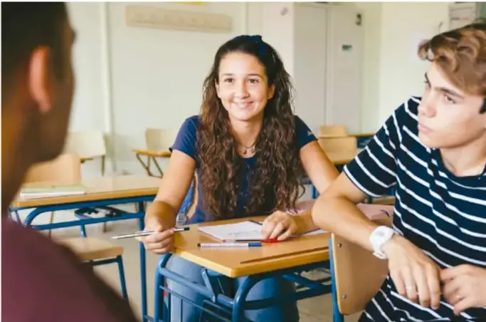 Tres estudiantes de secundaria sentados en pupitres de un salón de clase, conversando y tomando notas sobre una hoja; imagen de trabajo colaborativo.