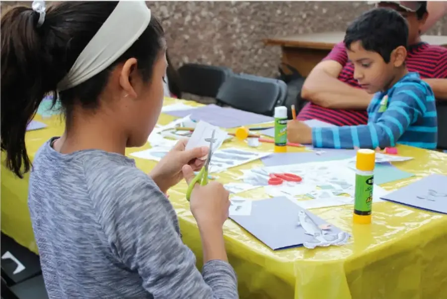 Fotografía de dos estudiantes de primaria sentados a una mesa con mantel amarillo, recortando y pegando imágenes para un collage; sobre la mesa hay tijeras, pegamento, hojas y recortes.