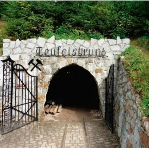 Fotografía de la entrada de una mina subterránea: portón de piedra con el nombre "Teufelsgrube" y un escudo con herramientas mineras (pico y martillo cruzados).