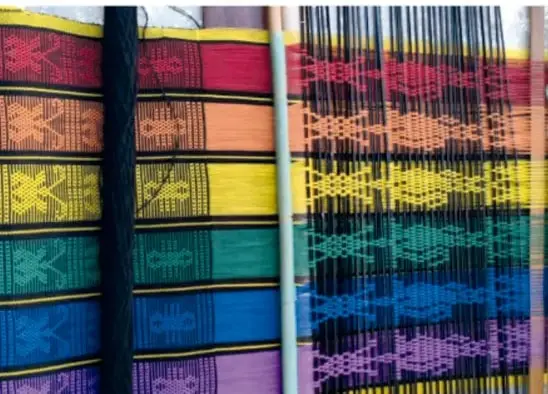 Mujer joven tejiendo en un telar de cintura una tela de muchos colores (rojo, azul, amarillo, morado) con diseños geométricos.