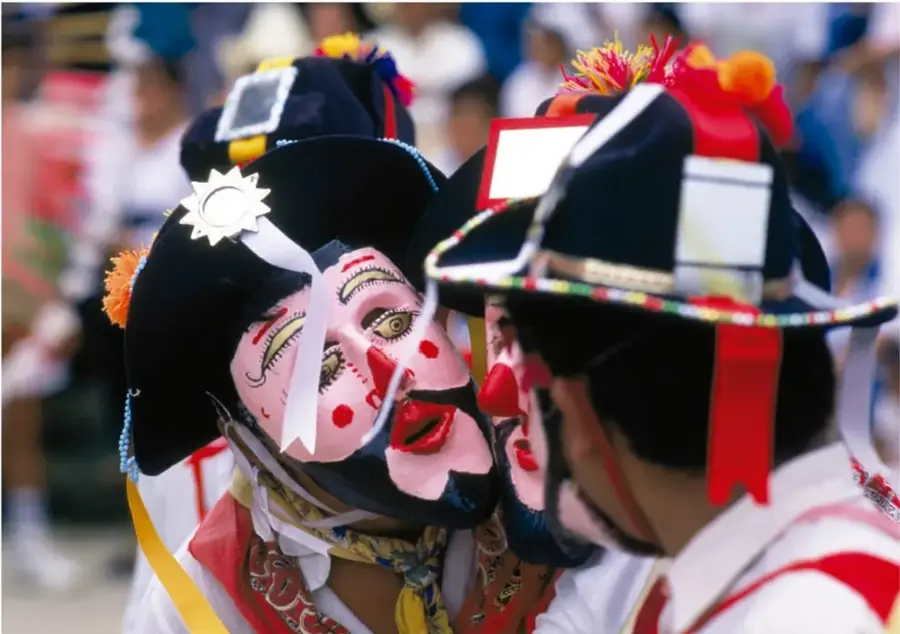 Dos danzantes frente a frente con máscaras rosadas de rasgos pintados, sombreros negros adornados con espejos, flores de papel naranja y listones; fondo de gente en la fiesta. Foto: Carlos Hahn, Ciudad de México.