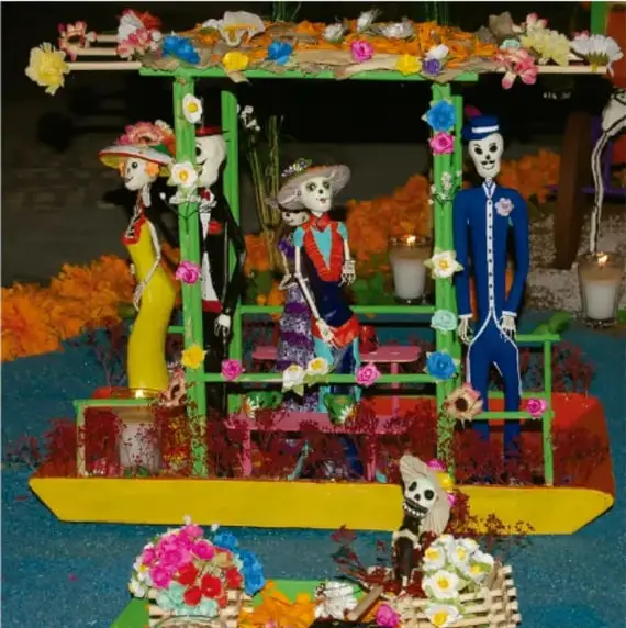 Artesanía de Día de Muertos con figuras de calaveras vestidas de época sobre una plataforma decorada con flores y papel picado.