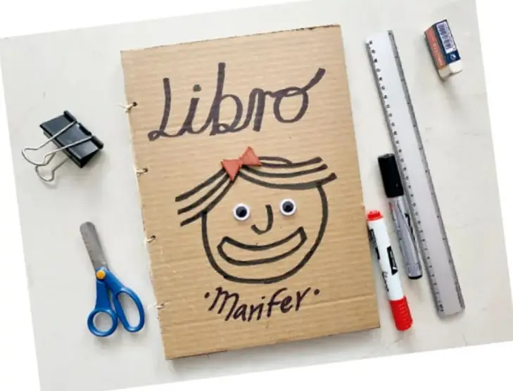 Fotografía de un libro artesanal elaborado con cartón, con la palabra 'Libro' escrita a mano, un dibujo de un rostro sonriente y el nombre 'Manfer'. Alrededor hay tijeras, clips, regla, marcadores y un borrador.