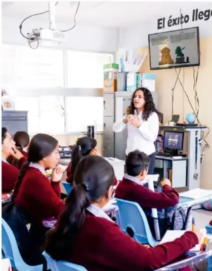 Aula con la maestra de pie frente al grupo presentando contenido en la pantalla; estudiantes en uniforme con el lema 'El éxito llega...' en la pared.