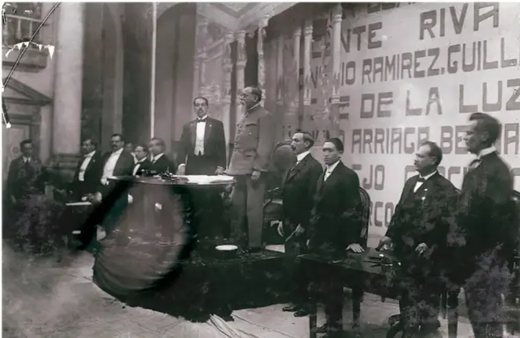 Fotografía en blanco y negro de la ceremonia de promulgación de la Constitución de 1917, con Venustiano Carranza y otros funcionarios en un salón con banderas y letreros alusivos.