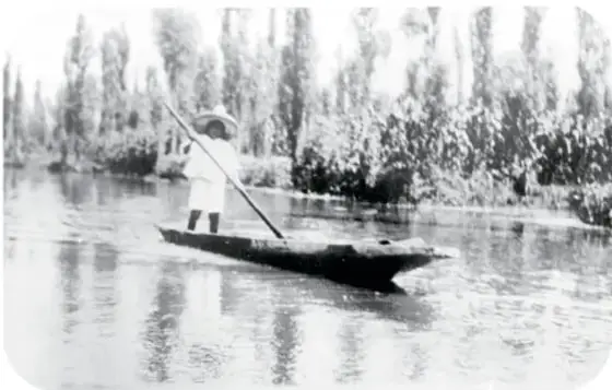 Fotografía antigua de una persona con sombrero de paja parada en una canoa o trajinera, remando en un canal rodeado de árboles.