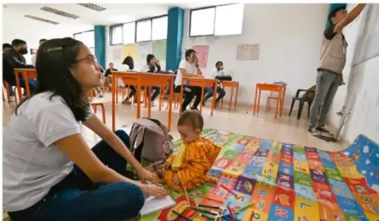 Fotografía de un aula con una docente sentada en el piso junto a una alfombra de letras, un infante pequeño y estudiantes trabajando en mesas al fondo.