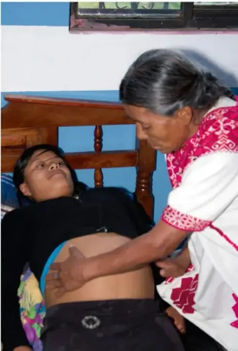 Fotografía de una partera indígena atendiendo a una mujer embarazada recostada en una cama; la partera palpa el vientre de la madre. La escena muestra la atención tradicional del embarazo y el parto.