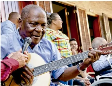 Fotografía de un hombre afrodescendiente mayor cantando y tocando la guitarra con un micrófono, acompañado por otras personas en un entorno comunitario al aire libre.