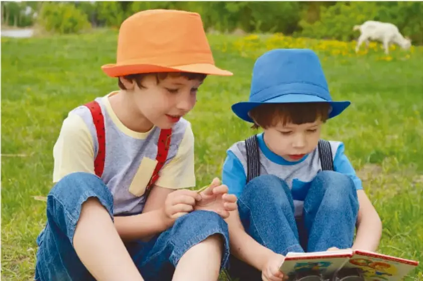 Dos niños sentados en el pasto leen un libro; al fondo hay un perro blanco. Ilustra la lectura compartida de poemas.