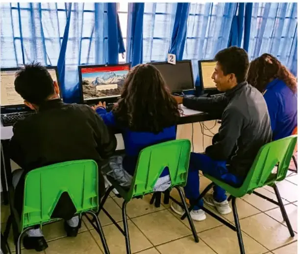 Fotografía de estudiantes trabajando en equipo frente a computadoras, organizados en grupos, con sillas verdes.