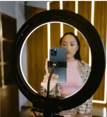 Fotografía de una joven grabándose con el teléfono celular frente a un anillo de luz (ring light), símbolo de la creación de contenido y la influencia de las redes sociales en la imagen personal.