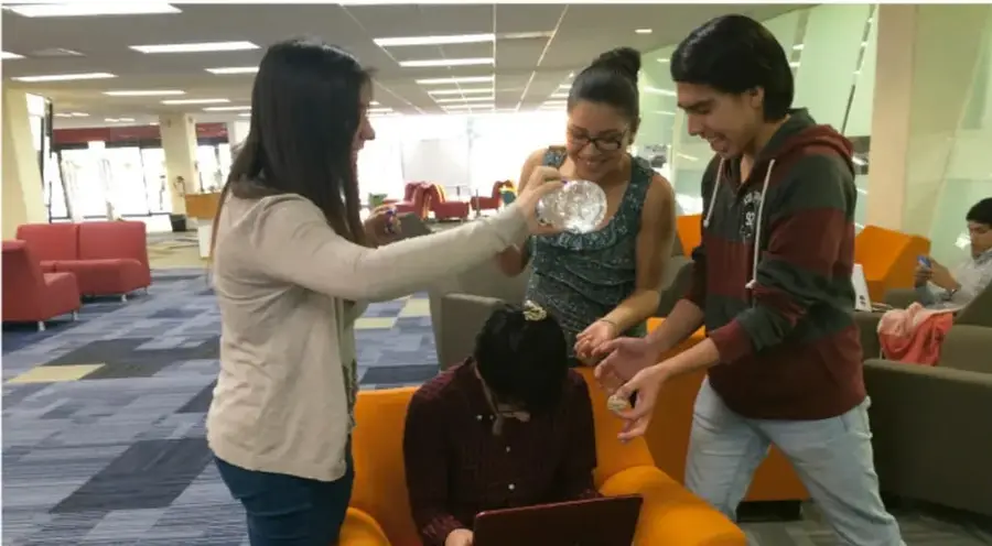 Fotografía de tres jóvenes en un espacio común (sillones naranjas y rojos, alfombra azul) interactuando alegremente; uno de ellos usa una computadora portátil.
