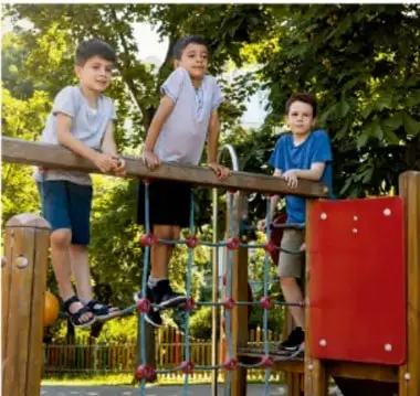 Fotografía de tres niños en un parque infantil. Dos niños están de pie sobre una estructura de madera y otro niño con camiseta azul está sentado en una barra, sonriendo. Al fondo hay árboles y vegetación.