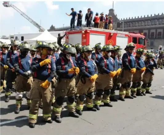 Fotografía de bomberos en fila con uniforme de trabajo amarillo y equipo de protección, frente a un camión de bomberos rojo; sobre el camión hay personas fotografiando; al fondo un edificio histórico con banderas (desfile cívico en el Zócalo de la Ciudad de México).