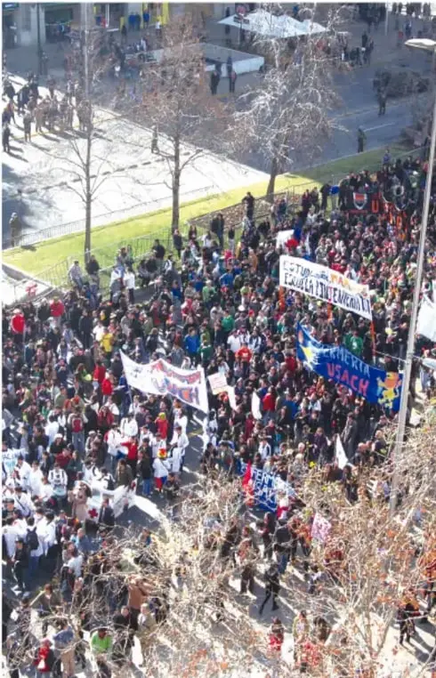 Multitud en una protesta en una calle arbolada, con pancartas y banderas.