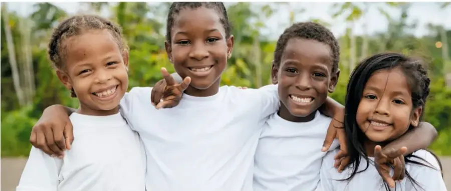 Fotografía de cuatro niños y niñas sonriendo, abrazados, al aire libre con fondo de vegetación.