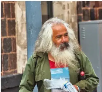 Fotografía de un hombre mayor con barba y cabello largo, sentado en la calle, sosteniendo un objeto. Ilustra la realidad de la desigualdad y la vulnerabilidad social.