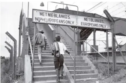 Fotografía histórica de una escalera de acceso a una estación con carteles bilingües de segregación racial: "NET NIE-BLANKES / NON-EUROPEANS ONLY" y "NET BLANKES / EUROPEANS ONLY", con personas ascendiendo por las gradas.