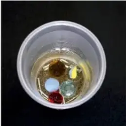Vaso visto desde arriba con la mezcla ya preparada: aceite, canicas y clavos, mientras la cuchara metálica la mezcla (pasos 2 y 3).