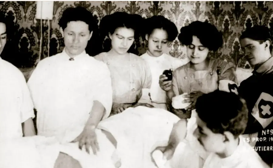 Fotografía histórica en blanco y negro de 1911: Elena Arizmendi y voluntarias de la Cruz Blanca Neutral impartiendo auxilios y curando heridos en Ciudad Juárez; se ven varias mujeres con víveres y vendajes junto a un herido y un hombre con brazalete de la Cruz.