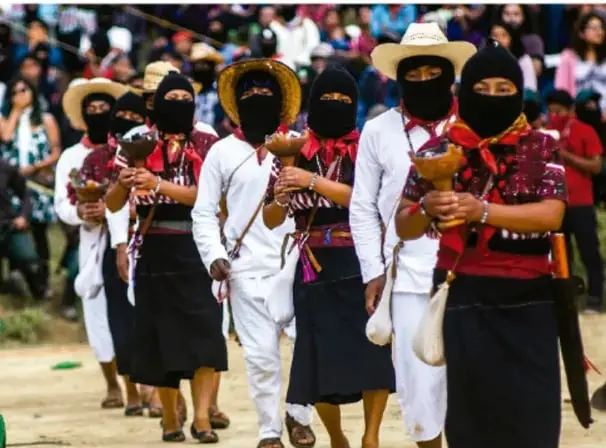 Fotografía de una comunidad indígena mexicana: mujeres con trajes tradicionales, huipiles y rebozos, rostros cubiertos con rebozo, cargando objetos rituales o cotidianos, acompañadas por hombres con sombrero, en un acto comunitario al aire libre.