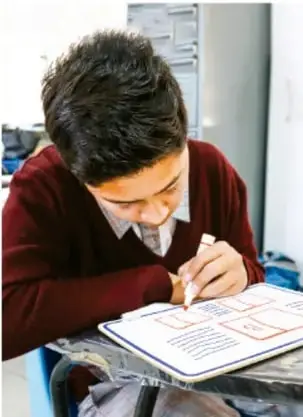 Fotografía de un estudiante que dibuja y colorea un boceto de texto ilustrado en una mesa.