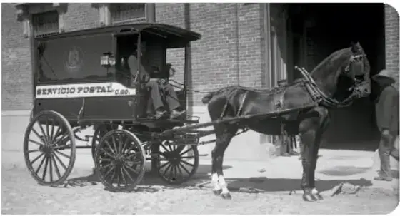 Fotografía en blanco y negro de un carruaje tirado por caballos con la leyenda 'Servicio Postal' en el costado, utilizado para transportar correo durante el Porfiriato.