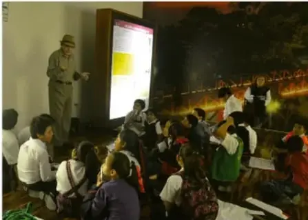 Fotografía de una actividad comunitaria al aire libre: un adulto con sombrero de paja frente a una pantalla o pizarrón iluminado, con un grupo de niñas, niños y jóvenes sentados en el suelo presenciando la presentación; al fondo se distingue un telón o paisaje nocturno.