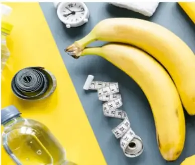 Fotografía de dos plátanos amarillos sobre fondo gris, con una cinta métrica enrollada, una botella de aceite y una bandeja amarilla, símbolo de medir la alimentación.