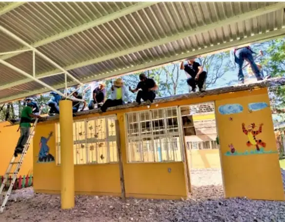 Fotografía de personas realizando trabajo comunitario bajo una estructura techada, probablemente reparación o construcción de un espacio público.