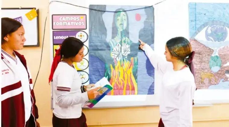 Tres estudiantes frente a un mural ilustrativo en el aula; una señala la ilustración mientras otra lee notas, junto a carteles de campos formativos.