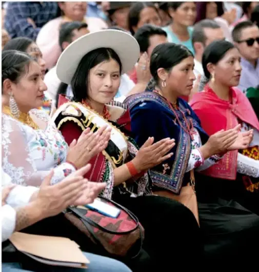 Fotografía de mujeres indígenas y mestizas con atuendos tradicionales aplaudiendo en un evento comunitario.