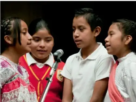 Fotografía de cuatro niños indígenas (dos niñas con vestimenta tradicional y dos niños con camisa blanca) frente a un micrófono, aparentemente participando en una presentación o discurso comunitario.