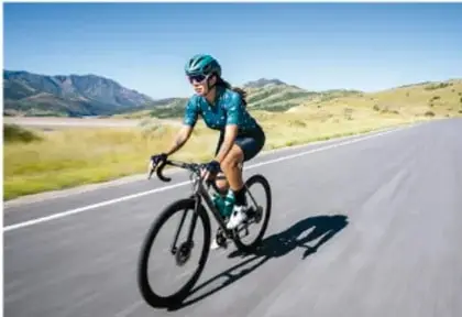 Fotografía de un ciclista con casco y ropa deportiva pedaleando por una carretera entre montañas, bajo un cielo despejado.