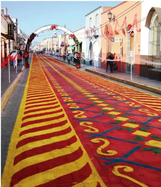 Fotografía de una calle alfombrada con motivos coloridos (rojo y amarillo) y arcos decorativos durante una celebración tradicional, probablemente una fiesta patronal mexicana.