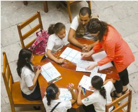 Fotografía de un grupo de estudiantes y una docente sentados alrededor de una mesa de madera, trabajando con hojas y cuadernos en un salón de clases.