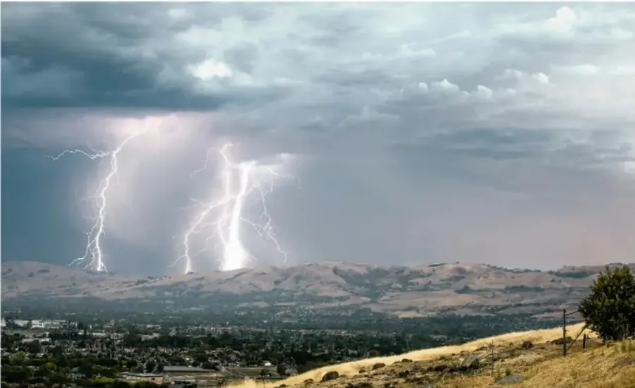 Fotografía de una tormenta eléctrica con varios rayos cayendo sobre un paisaje de colinas secas y una ciudad al fondo, que ilustra un riesgo natural.