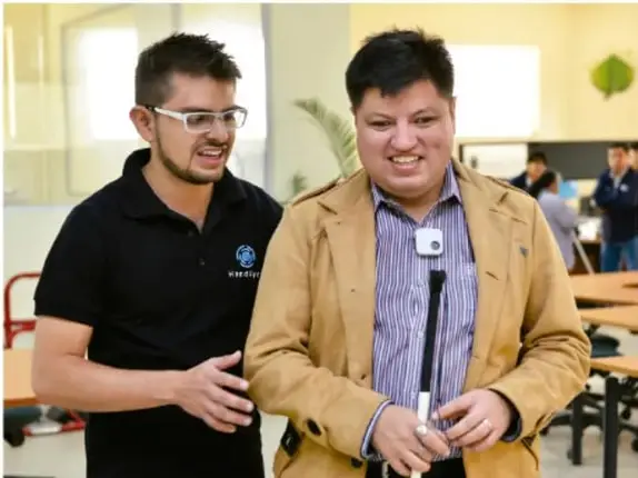 Fotografía de dos hombres en un espacio escolar; uno de ellos tiene discapacidad visual y usa bastón, mientras el otro lo guía con la mano.