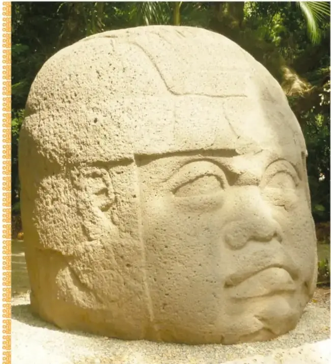 Fotografía a color de una cabeza colosal olmeca exhibida al aire libre, con su casco tallado y rasgos serenos, entre vegetación tropical.