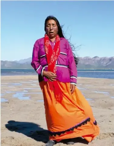 Fotografía de una mujer indígena con falda naranja, blusa rosa y rebozo rojo, de pie a la orilla de un lago en un paisaje montañoso, ilustrando el tema de las lenguas originarias.