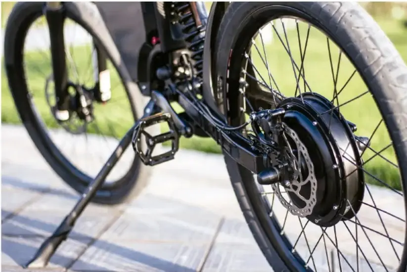 Fotografía de la parte trasera de una bicicleta negra con freno de disco, pedal y rueda con radios, sobre una superficie de piedra con césped al fondo.